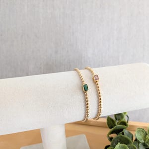 May include: Two gold beaded bracelets, each with a rectangular gemstone. One bracelet features a green gemstone, the other a pink gemstone. The bracelets are displayed on a white cylindrical stand against a neutral background.