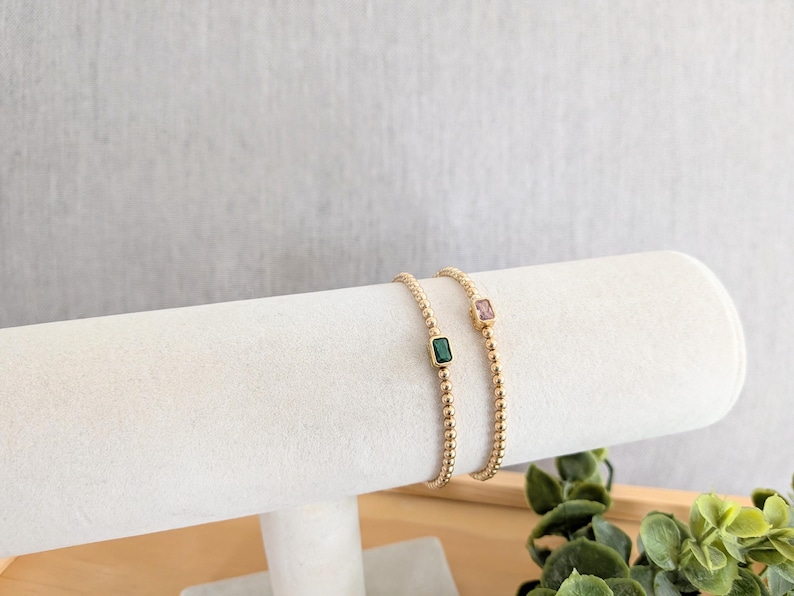 May include: Two gold beaded bracelets, each with a rectangular gemstone. One bracelet features a green gemstone, the other a pink gemstone. The bracelets are displayed on a white cylindrical stand against a neutral background.