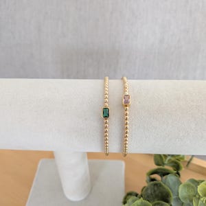 May include: Two gold beaded bracelets, each with a rectangular gemstone. One bracelet features a green gemstone, the other a pink gemstone. The bracelets are displayed on a white cylindrical jewelry holder.