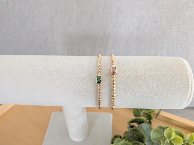 May include: Two gold beaded bracelets, each with a rectangular gemstone. One bracelet features a green gemstone, the other a pink gemstone. The bracelets are displayed on a white cylindrical jewelry holder.