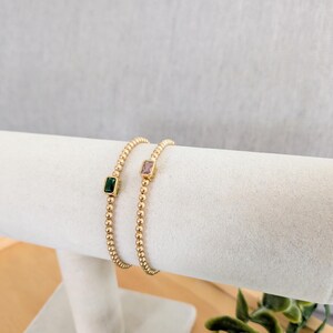 May include: Two gold beaded bracelets, each featuring a square gemstone. One bracelet has a green gemstone, the other a pink gemstone. The bracelets are displayed on a white, cylindrical jewelry holder.