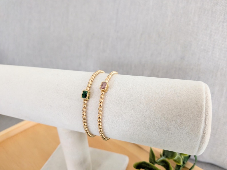 May include: Two gold beaded bracelets, each featuring a square gemstone. One bracelet has a green gemstone, the other a pink gemstone. The bracelets are displayed on a white, cylindrical jewelry holder.