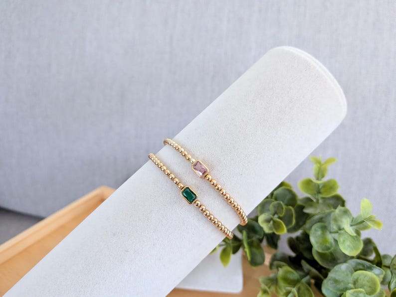 May include: Two gold beaded bracelets, each with a rectangular gemstone. One bracelet features a pink gemstone, the other a green gemstone. The bracelets are displayed on a white cylindrical surface.