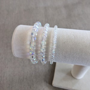 K&ouml;nnte beinhalten: Drei irisierende, klare Perlenarmb&auml;nder, die auf einem wei&szlig;en Ausstellungsst&auml;nder gestapelt sind. Die Armb&auml;nder bestehen aus kleinen, runden Perlen.