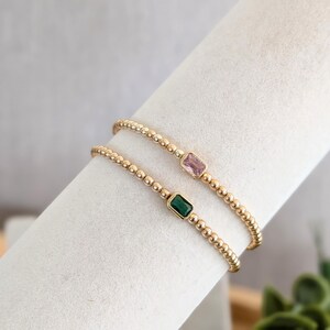 May include: Two gold beaded bracelets, one with a pink rectangular gemstone and the other with a green rectangular gemstone. The bracelets are displayed on a white surface.