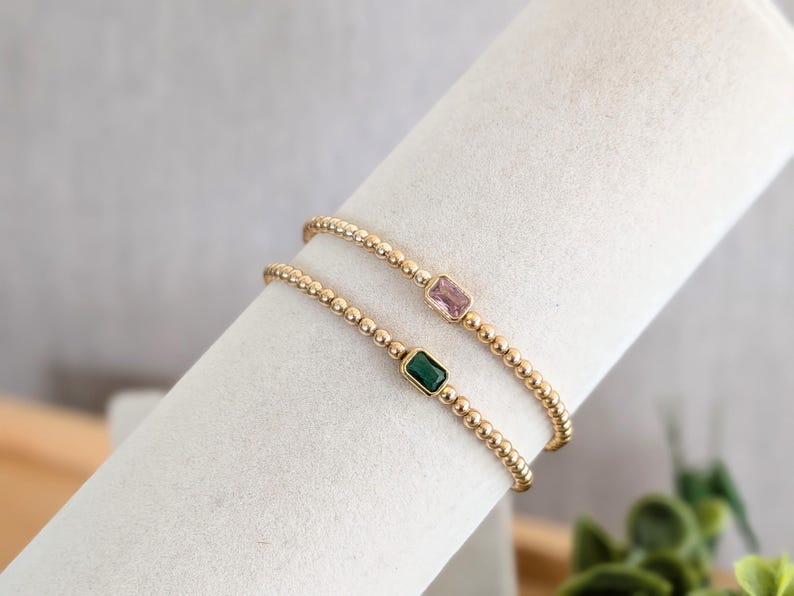 May include: Two gold beaded bracelets, one with a pink rectangular gemstone and the other with a green rectangular gemstone. The bracelets are displayed on a white surface.