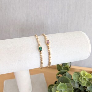 May include: Two gold beaded bracelets, each with a rectangular gemstone. One bracelet features a green gemstone, the other a pink gemstone. The bracelets are displayed on a white cylindrical stand, with greenery in the background.