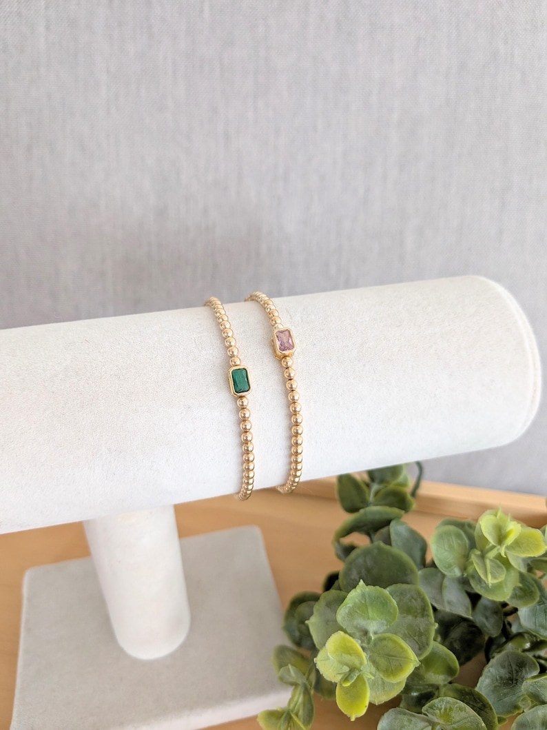 May include: Two gold beaded bracelets, each with a rectangular gemstone. One bracelet features a green gemstone, the other a pink gemstone. The bracelets are displayed on a white cylindrical stand, with greenery in the background.