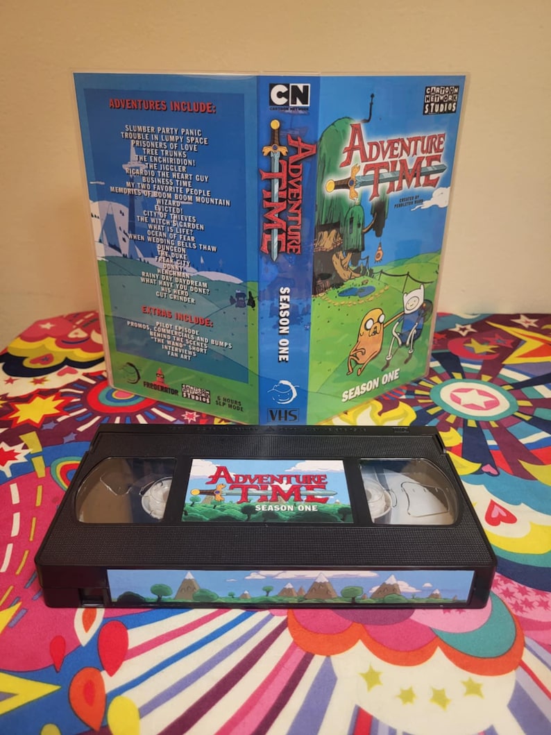 May include: A VHS tape and case for Adventure Time Season One. The case is blue with the show's logo and characters. The tape itself is black with a label featuring the show's title and characters. The case lists the episodes included.
