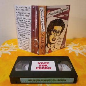 May include: A black VHS tape labeled "Napoleon Dynamite Collection" with a "Vote for Pedro" sticker. The case features a drawing of a man with glasses and the text "Napoleon Dynamite Collection". Additional text on the case describes the content.