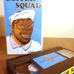 May include: A VHS tape and its cover featuring the text "LOITER SQUAD." The cover art depicts a cartoon portrait of a person wearing glasses and a head covering. The tape is black with a blue label.