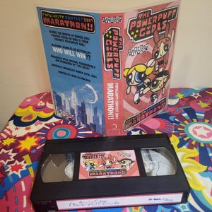 May include: A black VHS tape of "The Powerpuff Girls Marathon" with a pink and red cover. The cover features the Cartoon Network logo and the Powerpuff Girls characters. The text on the cover reads "Popularity Contest 2001 Marathon!!".