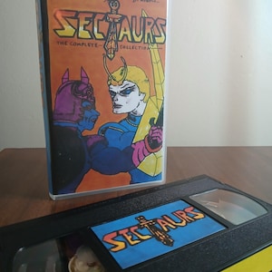 May include: A VHS tape and case for the animated series "Sectaurs: The Complete Collection." The case features a colorful illustration of two characters, with the show's title in large, stylized letters. The VHS tape is black with a yellow stripe.