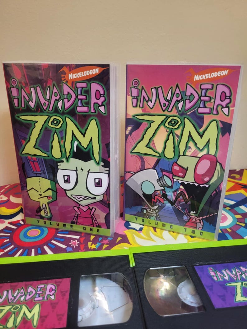 May include: Two Invader Zim VHS tapes and two DVD cases are displayed. The DVD cases are labeled "Volume One" and "Volume Two" with the Nickelodeon logo. The tapes and cases feature the show's title and characters in vibrant colors.