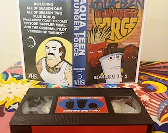May include: A VHS tape and case for the animated series Aqua Teen Hunger Force. The case features the show's logo and characters, with text indicating seasons one and two. The VHS tape itself has a red and black label with the show's title.