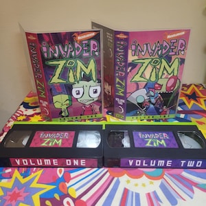 May include: Two VHS tapes and their cases for the animated series Invader Zim. The cases are labeled Volume One and Volume Two, with the Nickelodeon logo. The tapes are black with colorful labels featuring characters from the show.