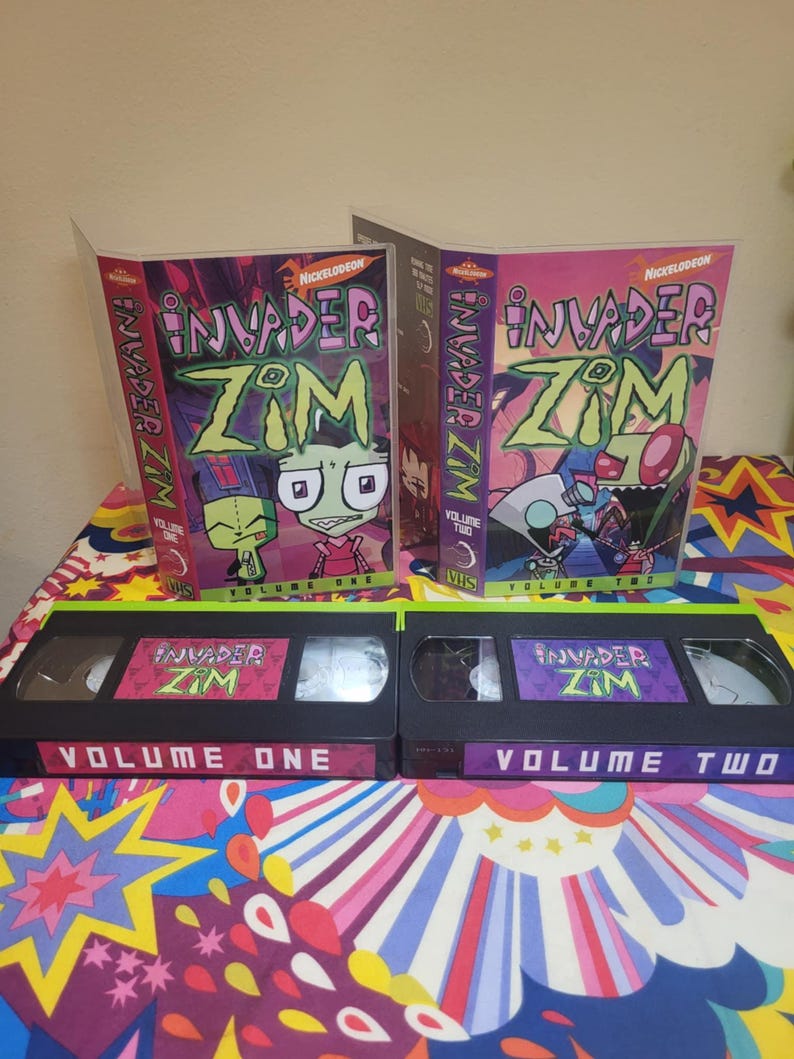 May include: Two VHS tapes and their cases for the animated series Invader Zim. The cases are labeled Volume One and Volume Two, with the Nickelodeon logo. The tapes are black with colorful labels featuring characters from the show.