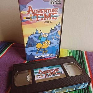 May include: A black VHS tape and DVD case for the animated series "Adventure Time: Season Three." The DVD case features the show's logo and characters Finn and Jake. The VHS tape has a matching label with the show's logo.