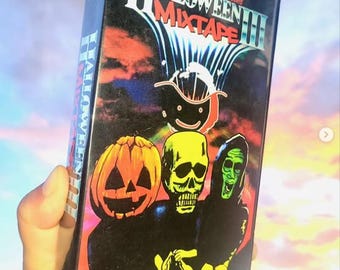 Może przedstawiać: Czarna kaseta VHS z napisem "Halloween Mixtape III". Okładka przedstawia dynię, czaszkę i zieloną postać. Kaseta jest trzymana na tle kolorowego nieba.