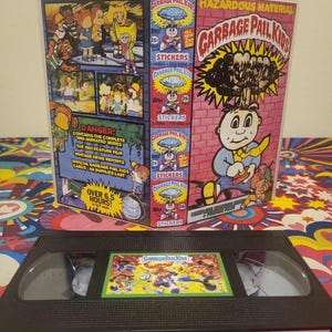 May include: A vintage VHS tape and box set featuring "Garbage Pail Kids" artwork. The box displays cartoon characters and the text "Hazardous Material." The VHS tape has a colorful label with the series' logo. The box also includes the text "Stickers" and "Over 6.5 Hours!"