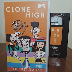 May include: A VHS tape and case for the animated series "Clone High." The case features cartoon characters against an orange background with the show's title. The VHS tape has the show's title on a label.
