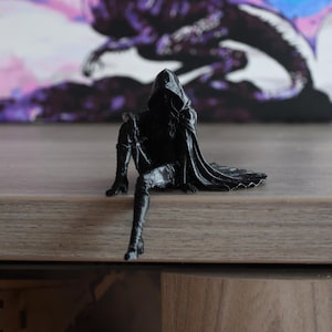 K&ouml;nnte beinhalten: Eine schwarze Figur einer Kapuzengestalt in sitzender Position mit einem nach unten ausgestreckten und einem angewinkelten Bein. Die Figur tr&auml;gt einen langen Umhang, der nach hinten f&auml;llt, hohe Stiefel und eine Kapuze, die das Gesicht verdeckt.