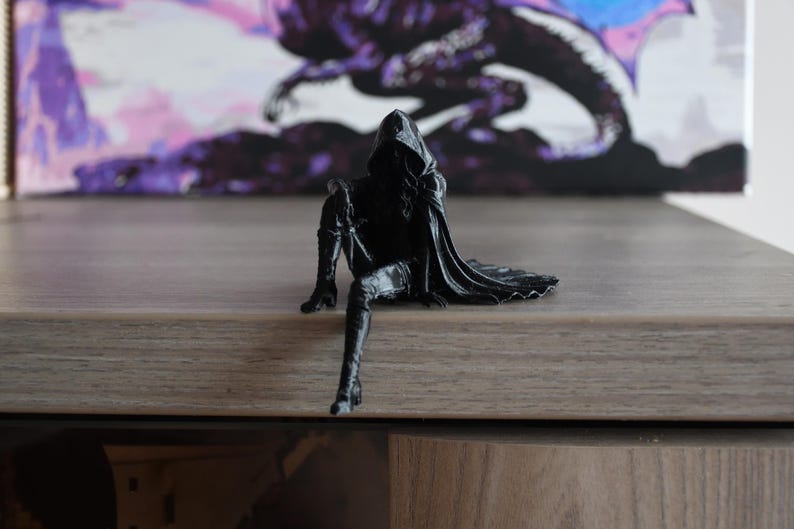 K&ouml;nnte beinhalten: Eine schwarze Figur einer Kapuzengestalt in sitzender Position mit einem nach unten ausgestreckten und einem angewinkelten Bein. Die Figur tr&auml;gt einen langen Umhang, der nach hinten f&auml;llt, hohe Stiefel und eine Kapuze, die das Gesicht verdeckt.