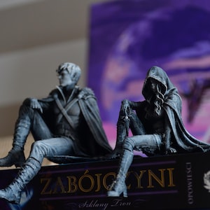 K&ouml;nnte beinhalten: Zwei detaillierte graue Figuren sitzen auf einem Buch mit dem Titel ZAB&Oacute;JCZYNI. Die linke Figur ist eine Person mit kurzem Haar und Umhang, w&auml;hrend die rechte Figur einen Kapuzenumhang tr&auml;gt. Beide Figuren sitzen in einer entspannten Haltung.