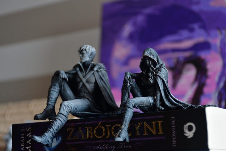 K&ouml;nnte beinhalten: Zwei detaillierte graue Figuren sitzen auf einem Buch mit dem Titel ZAB&Oacute;JCZYNI. Die linke Figur ist eine Person mit kurzem Haar und Umhang, w&auml;hrend die rechte Figur einen Kapuzenumhang tr&auml;gt. Beide Figuren sitzen in einer entspannten Haltung.