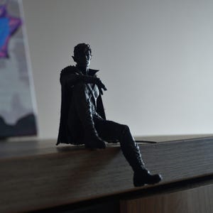 K&ouml;nnte beinhalten: Eine dunkle, silhouettenartige Figur eines sitzenden Charakters mit spitzen Ohren, der einen langen Mantel und Stiefel tr&auml;gt. Die Figur ist in einer entspannten Sitzposition mit einem ausgestreckten Bein dargestellt und zeigt detaillierte Texturen.