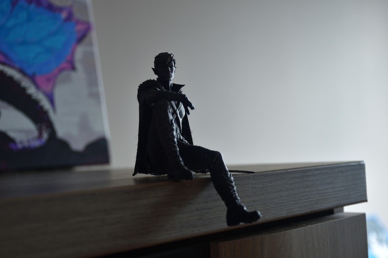 K&ouml;nnte beinhalten: Eine dunkle, silhouettenartige Figur eines sitzenden Charakters mit spitzen Ohren, der einen langen Mantel und Stiefel tr&auml;gt. Die Figur ist in einer entspannten Sitzposition mit einem ausgestreckten Bein dargestellt und zeigt detaillierte Texturen.
