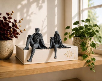 Op de afbeelding: Twee donkergrijze beeldjes van een persoon in een mantel en een persoon in een tuniek en cape, beide zittend op de rand van een groot, rechthoekig, gebroken wit boek. De beeldjes hebben gedetailleerde kledingtexturen, inclusief laarzen.