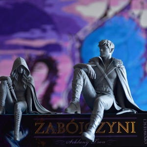 K&ouml;nnte beinhalten: Zwei graue Harzfiguren von Fantasy-Charakteren, die auf einem Buch mit dem Titel ZAB&Oacute;JCZYNI sitzen. Die linke Figur tr&auml;gt einen Kapuzenumhang und h&auml;lt einen Dolch. Die rechte Figur tr&auml;gt einen Umhang und Stiefel. Der Buchr&uuml;cken nennt Sarah J. Maas.