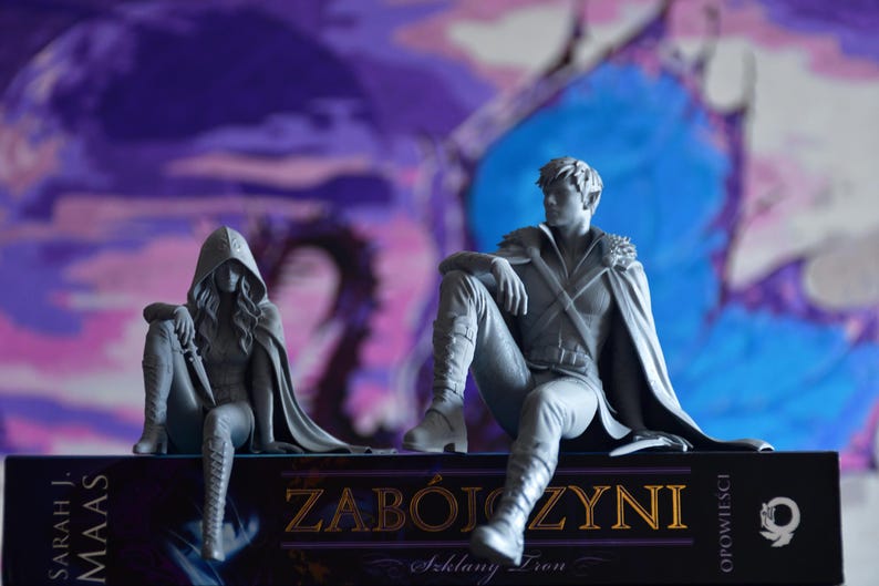 K&ouml;nnte beinhalten: Zwei graue Harzfiguren von Fantasy-Charakteren, die auf einem Buch mit dem Titel ZAB&Oacute;JCZYNI sitzen. Die linke Figur tr&auml;gt einen Kapuzenumhang und h&auml;lt einen Dolch. Die rechte Figur tr&auml;gt einen Umhang und Stiefel. Der Buchr&uuml;cken nennt Sarah J. Maas.