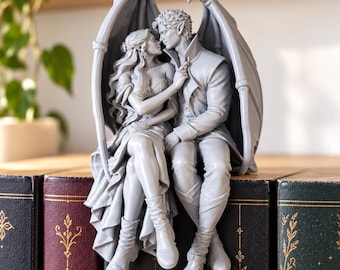 Op de afbeelding: Een grijs monochroom harsbeeldje van twee gevleugelde figuren met puntige oren die dicht bij elkaar zitten. De figuren zijn omhelsd, waarbij de een de kraag van de ander aanraakt, omlijst door grote, gebogen, leerachtige vleugels.