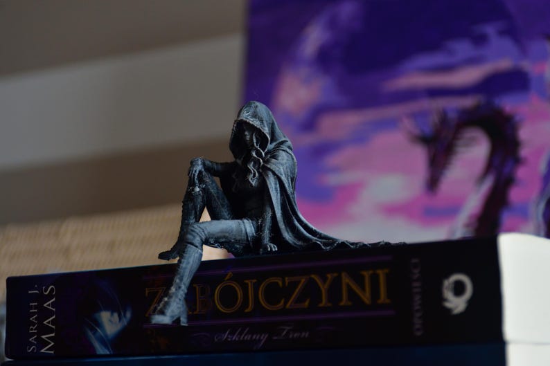 K&ouml;nnte beinhalten: Eine dunkelgraue Figur einer Kapuzengestalt, die auf dem Buchr&uuml;cken des Buches Zabojczyni von Sarah J. Maas sitzt. Die Figur tr&auml;gt einen langen Umhang und hohe Stiefel, ein Bein ist am Knie angewinkelt. Der Buchr&uuml;cken zeigt goldene Schrift und ein Logo.