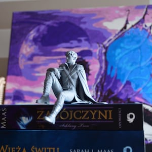 K&ouml;nnte beinhalten: Eine graue, unbemalte Harzfigur eines sitzenden m&auml;nnlichen Charakters mit spitzen Ohren, der einen Umhang und Stiefel tr&auml;gt, platziert auf einem Buch mit dem Titel "Zab&oacute;jczyni" von Sarah J. Maas, das auf einem Buch namens "Wieża Świtu" liegt.