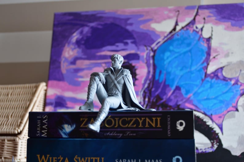 K&ouml;nnte beinhalten: Eine graue, unbemalte Harzfigur eines sitzenden m&auml;nnlichen Charakters mit spitzen Ohren, der einen Umhang und Stiefel tr&auml;gt, platziert auf einem Buch mit dem Titel "Zab&oacute;jczyni" von Sarah J. Maas, das auf einem Buch namens "Wieża Świtu" liegt.