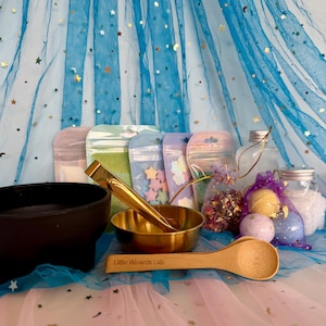 May include: An assortment of apothecary supplies, featuring a black bowl, a gold-coloured bowl, tongs, and a wooden spoon with the text "Little Wizards Lab." Several small bags and jars are also visible, set against a backdrop of blue tulle with gold stars.