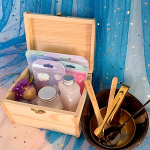 May include: A wooden box with a hinged lid, containing small bottles, pouches, and a purple decorative item. A coconut shell bowl holds spoons and tongs. The background is a blue fabric.