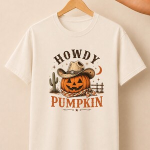 May include: A cream-colored short-sleeve t-shirt featuring a graphic of an orange jack-o'-lantern wearing a cowboy hat and a bandana. The text "HOWDY PUMPKIN" is printed above and below the pumpkin, accompanied by a cactus and fence.