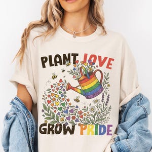 May include: An ivory t-shirt featuring a graphic of a rainbow-colored watering can pouring out colorful hearts and flowers. The text reads "PLANT LOVE GROW PRIDE" in black and rainbow lettering, accompanied by small bees and floral illustrations.