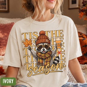 May include: An ivory short-sleeve t-shirt featuring a graphic of a raccoon wearing a brown knit hat, heart-shaped sunglasses, and a plaid scarf. The raccoon holds a maple leaf and a coffee cup. Text reads: TIS THE SEASON.