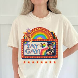 Puede incluir: Una camiseta de color marfil con un gráfico de marquesina de teatro con un arcoíris, el texto "IT'S OK TO SAY GAY!", un icono de corazón y la ilustración de una persona con sombrero de copa y gafas de sol arcoíris señalando hacia adelante, con estrellas rojas.