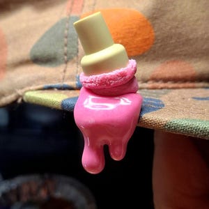 May include: A decorative pin attached to a tan fabric hat with a camouflage pattern. The pin features a pale yellow cylindrical top, a textured pink middle section, and a smooth, glossy pink base that appears to be melting downward.