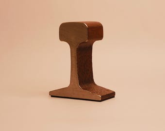 May include: A metallic object shaped like a cross-section of a railway track. The top and base have a smooth, polished finish, while the vertical web section features a textured, dark brown surface. A thin black base layer is visible at the bottom.