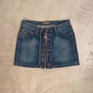 May include: A blue denim mini skirt featuring a central silver-toned metal zipper closure and a row of matching metal buttons along the front. The waistband includes a small rectangular label with the word Crown printed in a dark font.