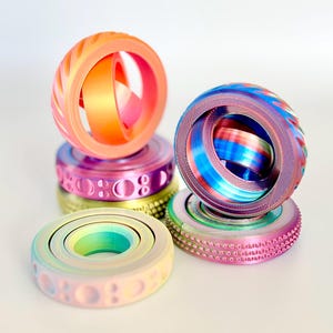 May include: A collection of colorful, 3D-printed fidget toys. The toys feature a ring-like design with inner rotating components. The color schemes include gradients of orange, pink, purple, blue, and green. The toys are stacked on top of each other.
