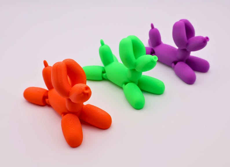 May include: Three colorful balloon dog sculptures in orange, green, and purple are arranged on a white surface. The dogs are in a playful pose, with their legs and tails extended. The sculptures are made of a smooth material.