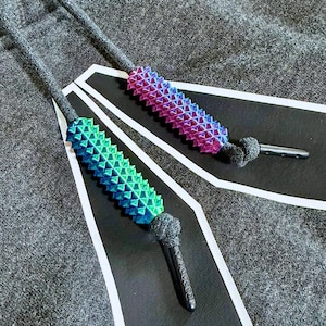 May include: Close-up of two shoelace tips with textured, geometric designs. One is green and blue, the other purple and blue. The tips are attached to gray cords and black tags with white stripes. The Reebok logo is visible on one tip.